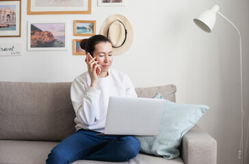 Wall Mural - Young woman working on laptop. Millennial girl working remote job from home, using mobile phone, sitting on the cozy sofa, smiling. 