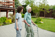 © pressmaster - Young caregiver supporting hand of senior patient of retirement home using walker during stroll in the garden with green lawns and trees