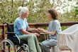 © pressmaster - Side view of young caregiver holding hands of senior female with disability sitting in wheelchair and listening to her on terrace of retirement home