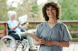 © pressmaster - Happy young nurse or caregiver with clipboard looking at camera with smile while standing against senior female patient of retirement home
