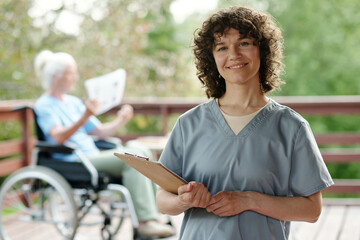 Happy young nurse or caregiver with clipboard looking at camera with smile while standing against senior female patient of retirement home