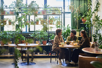 Group of young intercultural friends sitting by table in the corner of cafe with variety of green plants, having chat and coffee with snacks