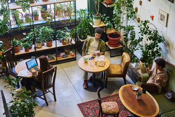 Poster - Three young intercultural coworkers using gadgets and looking through papers while sitting by tables and on couch in cafe with green plants