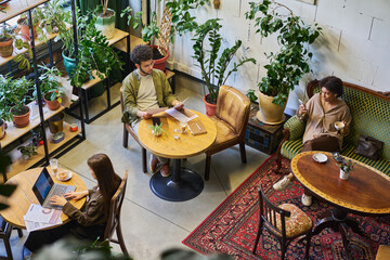 Wall Mural - Group of young intercultural analysts or brokers working in cafe while sitting by tables and on couch, using gadgets and looking through papers