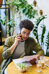 Poster - Young economist or broker talking on mobile phone and reading financial document while sitting by table in front of camera against green plants
