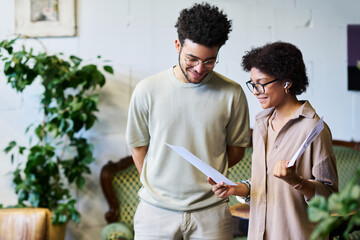 Leinwandbilder - Two happy young intercultural brokers in casualwear discussing financial documents while standing in coworking space with green plants