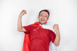 Excited Asian man wearing a red top, flag cape and headband, showing strong gesture by lifting his arms and muscles smiling proudly. Indonesia's independence day concept.