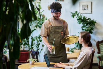 Aufkleber - Young waiter with glass of refreshing lemonade looking at pretty female guest of cafe sitting by table in front of laptop and analyzing data
