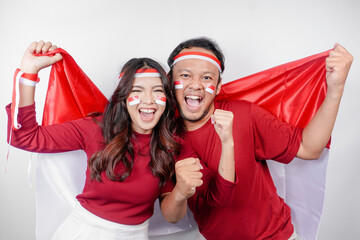 A young Asian couple with a happy successful expression wearing red top and headband while holding Indonesia's flag, isolated by white background. Indonesia's independence day concept.