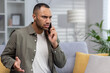 © Tetiana - Angry young African American man talking on the phone at home. Raises hands, solves problems