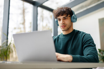 E-learning concept. Focused european guy in headphones looking at laptop, watching online lecture, sitting in classrom