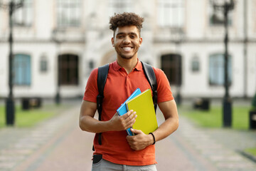 Wall Mural - Cheerful Black Student Guy Holding Workbooks Posing With Backpack Near College Building