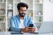 © Prostock-studio - Handsome young indian male using smartphone while sitting at desk at home