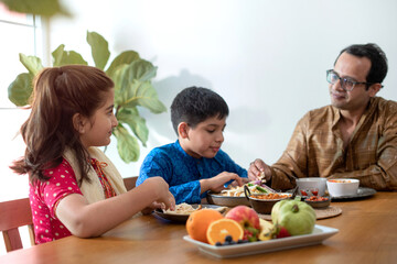 Wall Mural - Happy Indian father with daughter and son having meal at home, family in traditional dress tasting Indian food, selective focus