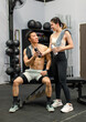 Asian strong young male muscular shirtless fitness model in sporty shorts and female athlete in sport bra sitting smiling taking break holding water bottle talking together in gym