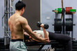 Asian strong young shirtless male muscular fitness trainer in sporty shorts standing helping saving female athlete in sexy sport bra lifting up metal dumbbells lay down on bench working out in gym.