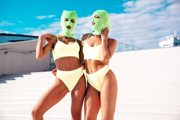 Two beautiful sexy women in green underwear. Models wearing bandit balaclava mask. Hot seductive female in nice lingerie posing in the street at sunny day, blue sky. Crime and violence. Perfect body