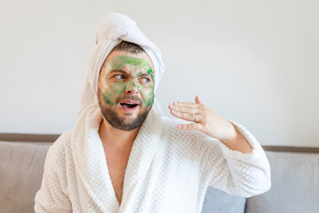 Surprised guy spends hours treating and caring for his skin, applies green face mask to pamper himself, wearing a soft bathrobe and a towel on his head