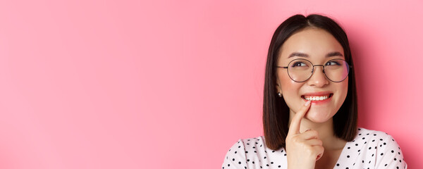 Headshot of cute asian woman in trendy glasses smiling, having an idea, thinking and looking at upper left corner, pink background