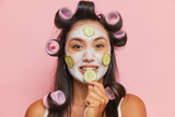 Fototapeta  - Face portrait of cute girl with curlers on long black hair, cosmetic procedures, cream mask with cucumbers, face treatment concept, copy space