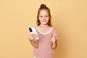 Wall Mural - Cute optimistic smiling little brown haired girl wearing striped t-shirt standing isolated over beige background holding smartphone browsing internet showing thumb up.