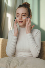 Wall Mural - cheerful woman closing eyes while applying face cream for healthy soft skin, sitting on comfortable 