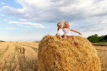 Two Cute Adorable Caucasian Siblings Enjoy Having Fun Sitting On Top Over Golden Hay Bale On Wheat Harvested Field Near Farm. Happy Childhood And Freedom Concept. Rural Countryside Scenic Landscape