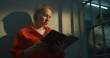 Female Prisoner In Orange Uniform Sits On Bed In Prison Cell, Reads Bible. Prison Officer Walks The Corridor. Woman Criminal Serves Imprisonment Term For Crime In Jail Or Correctional Facility.