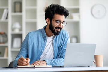 online lesson. smiling young indian man using laptop and taking notes