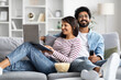 © Prostock-studio - Relaxed eastern couple resting at home, watching tv, using laptop