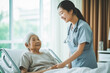 © Bobboz - Smiling Asian caring nurse supporting holding hand of olde senior female patient lying on bed at clinic or hospital. Elderly people health care concept. Generative AI.