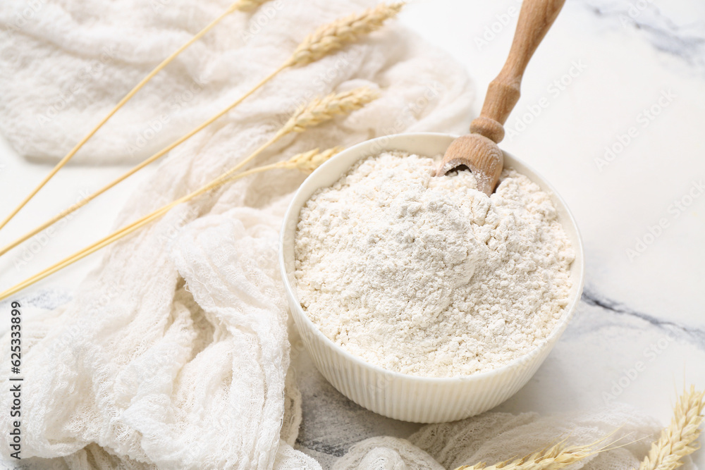 Bowl with wheat flour and scoop on white marble table, closeup