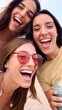 © CarlosBarquero - Vertical of Three excited young Caucasian friends in summer clothes taking selfie on beach. Group of smiling women enjoying vacation. Cheerful beautiful girls of generation z pose for photo with phone