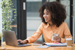 © Songsak C - Portrait of a beautiful confident businesswoman using a laptop computer holding a mobile phone sitting in a modern office. Smiling African American freelancer working online from home.