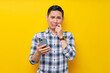 © Bangun Stock Photo - Worried young Asian man wearing a white checkered shirt holding smartphone and biting his nails, looking camera with a frustrated expression isolated over yellow background. People Lifestyle Concept