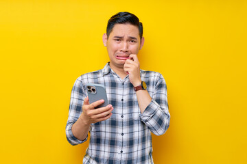 Worried young Asian man wearing a white checkered shirt holding smartphone and biting his nails, looking camera with a frustrated expression isolated over yellow background. People Lifestyle Concept