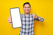 © Bangun Stock Photo - Smiling handsome young Asian man wearing a white checkered shirt showing a mobile phone with a blank screen and thumbs up sign isolated over yellow background. People Lifestyle Concept