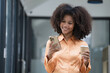 © Songsak C - Portrait of an African American businesswoman standing and holding a coffee cup and using a mobile phone, standing and smiling at an office.