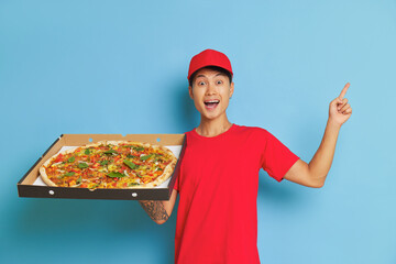 Wall Mural - Welcoming delivery man in red t-shirt and red cap on blue backdrop with large pizza in hand, points his index finger up with smile, food delivery concept, copy space