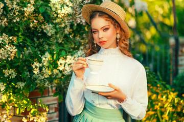 Art Happy red hair woman holding porcelain white cup in hands fantasy retro lady drinking tea, girl princess having tea beauty romantic face. summer garden tree park vintage style straw hat on head