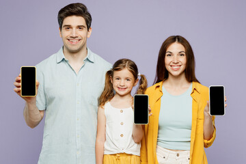 Wall Mural - Young parents mom dad with child kid daughter girl 6 years old wearing blue yellow casual clothes hold use blank screen area mobile cell phone isolated on plain purple background. Family day concept.