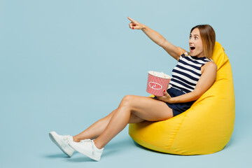 Full body young woman she wearing striped tank shirt casual clothes sit in bag chair eat popcorn point index finger aside on area isolated on plain pastel light blue cyan background studio portrait.