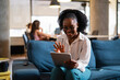 © JustLife - Beautiful African businesswoman sitting on sofa in office. Young woman using digital tablet.