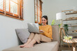 © Natee Meepian - Beautiful attractive young Asian woman sitting on gray sofa and using laptop at living room in the morning