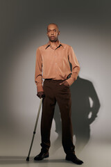 Wall Mural - real people, bold african american man with myasthenia gravis standing with walking cane on grey background, hand in pocket, pose, dark skinned person in shirt, diversity and inclusion, full length