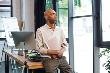 Wall Mural - dark skinned businessman with myasthenia disease holding folder and standing near desk and walking cane, bold african american office worker with ptosis eye syndrome, inclusion, monitor and graphics