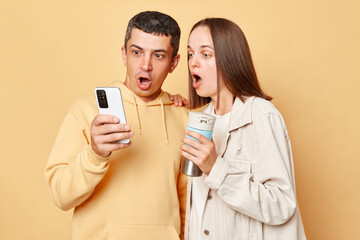 Wall Mural - Surprised beautiful woman and handsome man wearing casual style clothing standing isolated over beige background browsing internet together on smartphone drinking hot coffee from thermos.