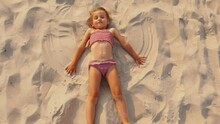 A Child Playing In A Brown Swimsuit Lying On The Beach On The Sand Makes An Snow Angel