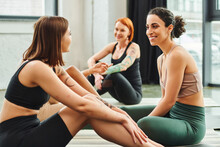 Young And Tattooed Woman In Sportswear Pointing With Finger And Talking To Multiracial Girlfriend During Yoga Class In Gym, Friendship, Harmony And Mental Health Concept