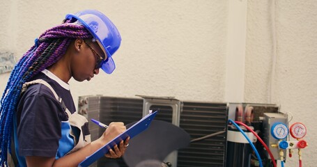 Wall Mural - Skilled african american expert commissioned for outdoor air conditioner annual check up, writing findings on clipboard. Seasoned wireman doing hvac system investigation of damaged parts